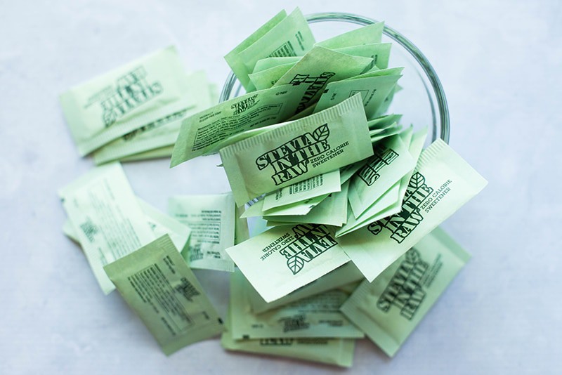 Overhead image of a bowl of stevia packets with many of the green colored packets overflowing around the bowl.