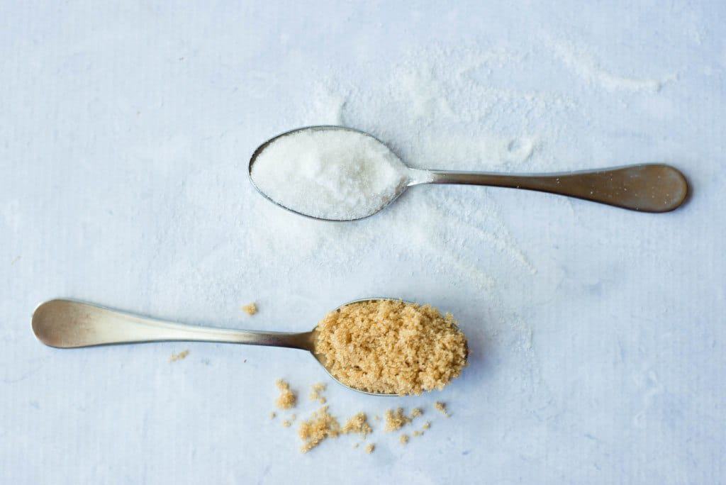 Overhead view of two spoons, one holding white refined sugar and the other brown refined sugar.