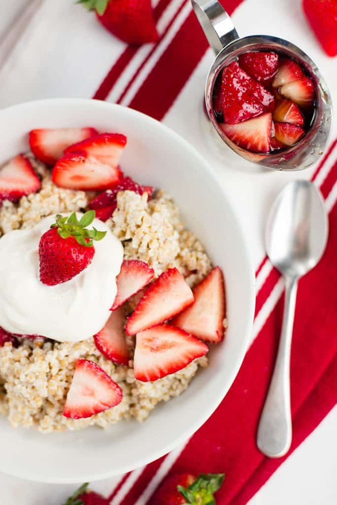 Overhead image of a white bowl filled with steel cut oats topped with strawberries and cream with a spoon and small carafe of strawberries off to the side.