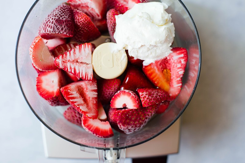 Frozen halved strawberries together with Greek yogurt placed in a blender to make strawberry frozen yogurt 