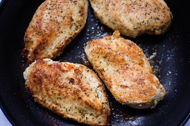 Four pieces of chicken breast covered in healthy almond meal breading and cooking in a skillet, ready to be baked to make the healthy chicken Parmesan dish 