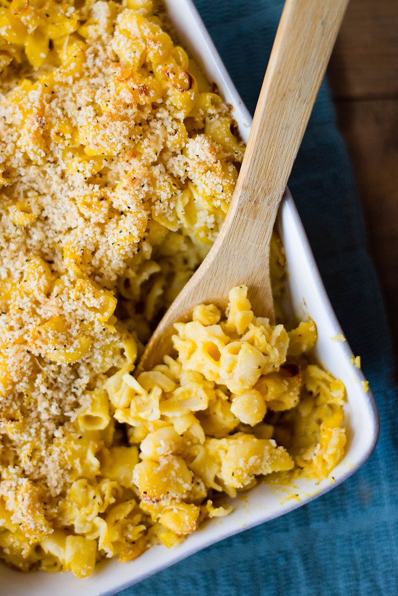 Close up view of a casserole dish of Healthy Mac And Cheese, with a wooden serving utensil dipping into the dish.