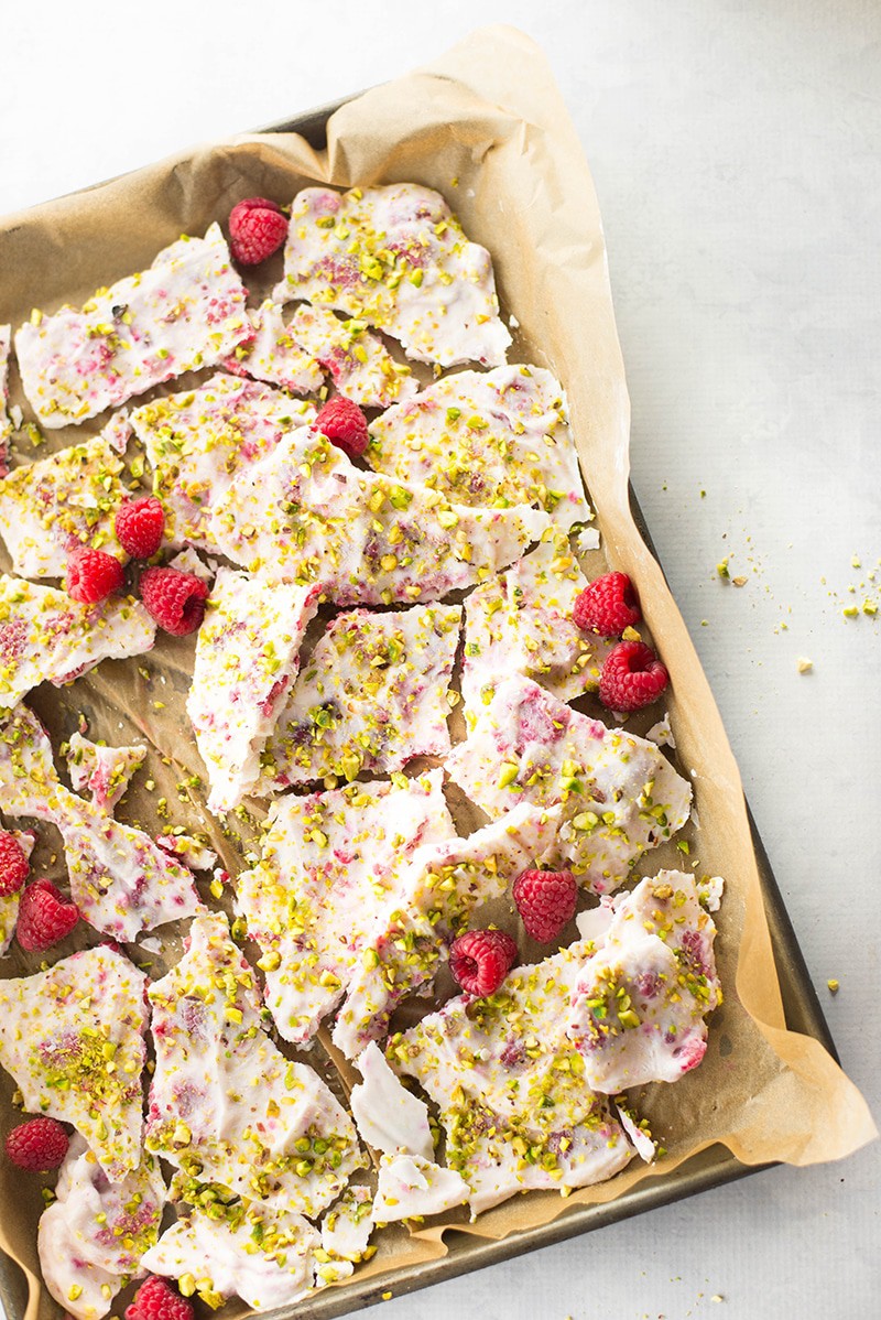 Overhead image of a sheet pan lined with parchment paper, containing broken pieces of Frozen Yogurt Bark With Raspberries & Pistachios.