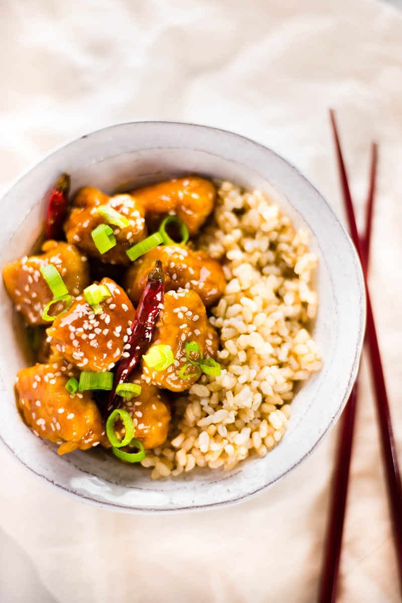 Overhead image of a white bowl containing Healthy General Tso's chicken, topped with sesame seeds and green onion, and with chop sticks beside the bowl.