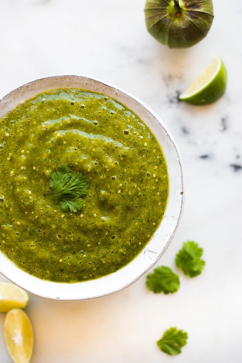 Overhead view of a bowl of prepared homemade tomatillo salsa verde, topped with a sprig of cilantro and a lime and tomatillo beside it.