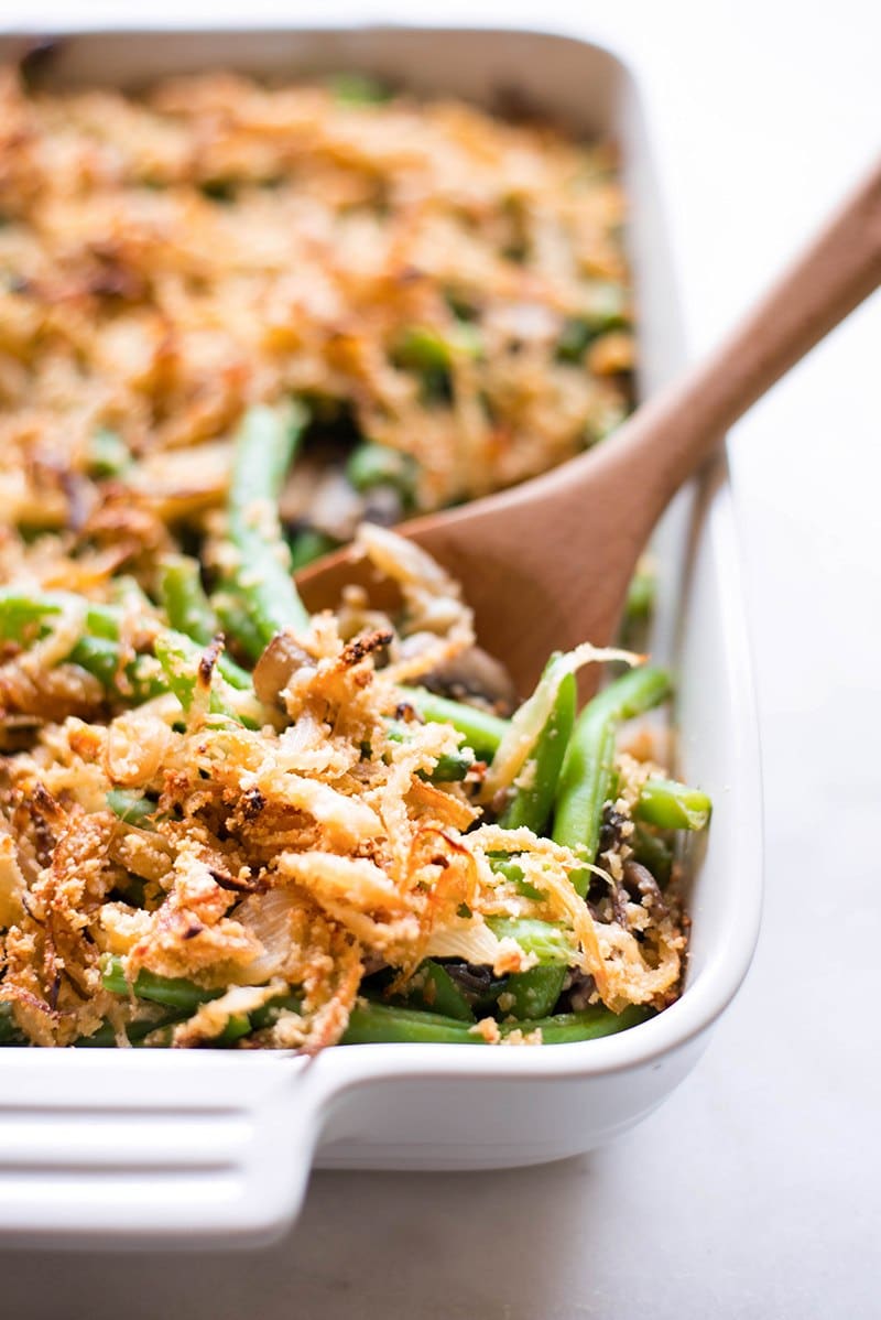 Overhead image of a white casserole dish filled with Healthy Green Bean Casserole, topped with crispy onions. A wooden spoon is dipped into the casserole.