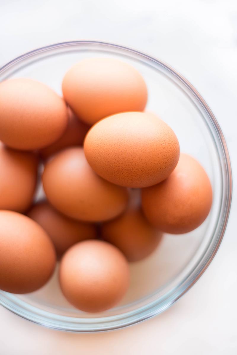 Overhead close up view of a clear glass bowl filled with uncooked brown eggs in the shell.