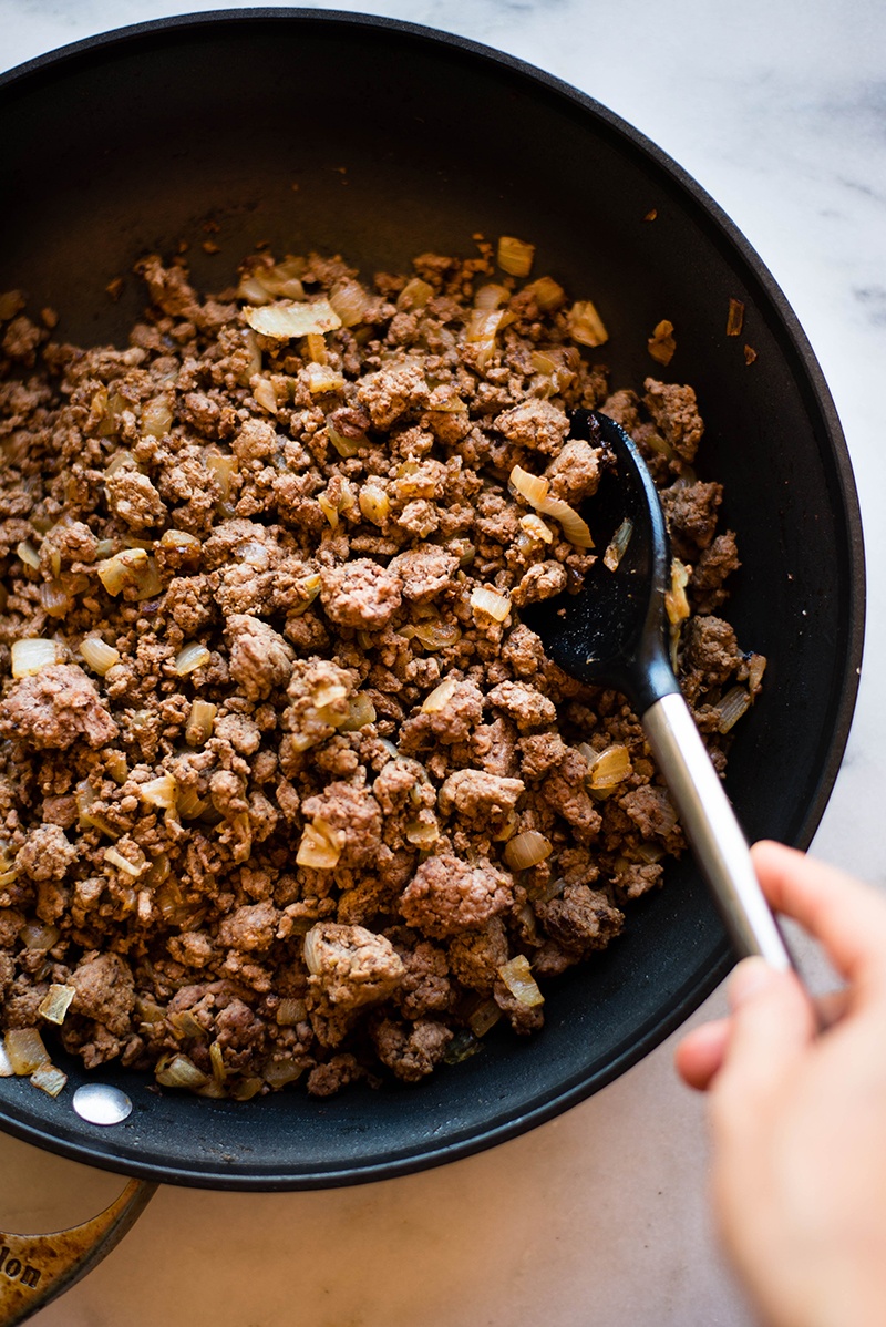 A skillet of ground buffalo being cooked for the Quinoa Taco Bowl; a hand holding a spoon is also in the image.