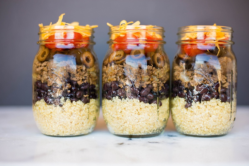 Side view of three mason jars containing Taco Stacker recipe, including ground meat, quinoa, black beans, olives, cheese and tomatoes.