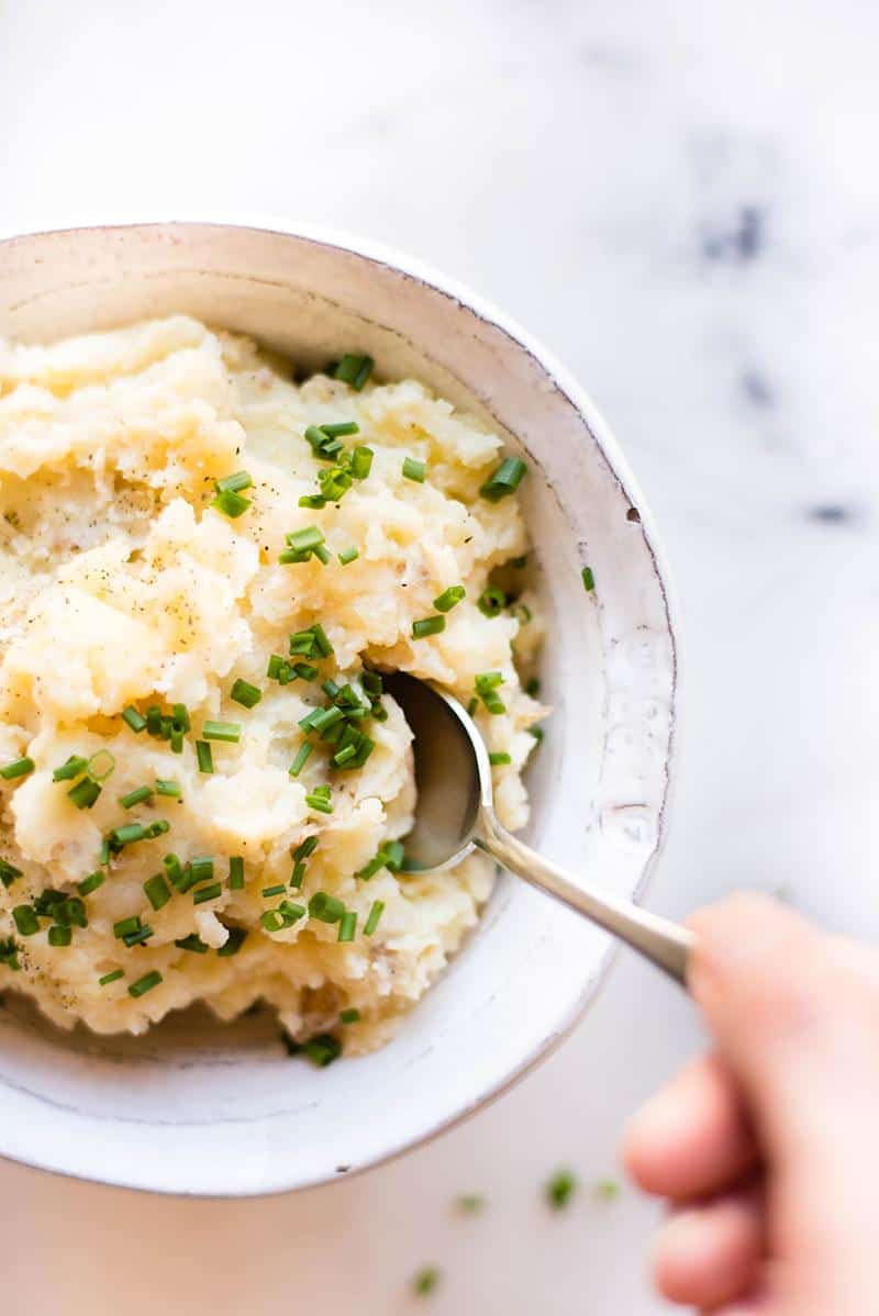 Overhead view of a white bowl filled with Crock Pot Mashed Potatoes garnished with chives.. A hand holding a spoon is dipping into the bowl.