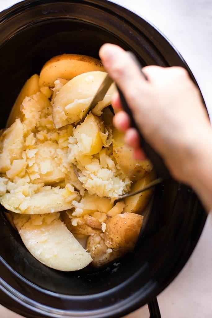Overhead view of a hand in motion mashing potatoes in a black crock pot.