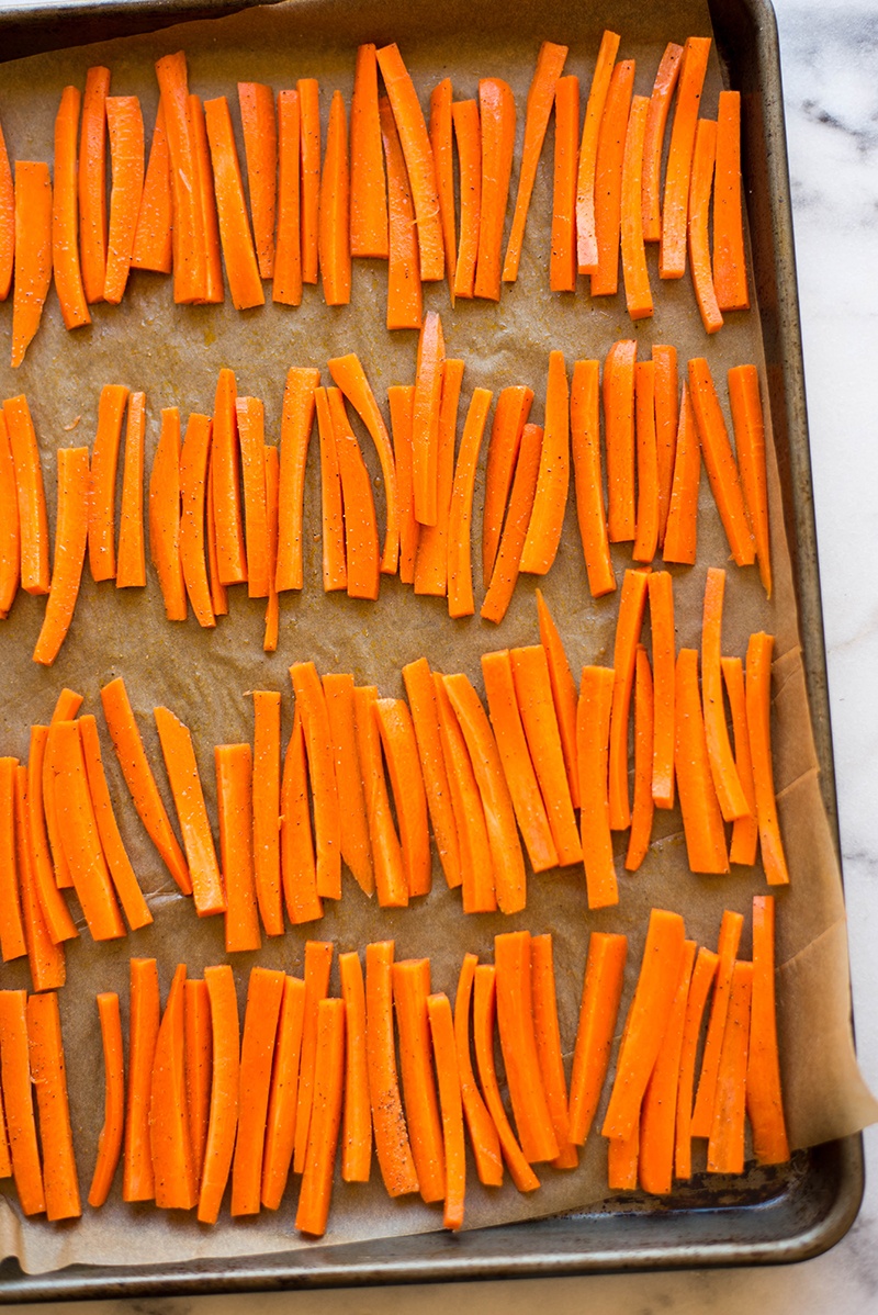Seasoned, raw carrot slices placed on a baking sheet, ready to be baked to make carrot fries.
