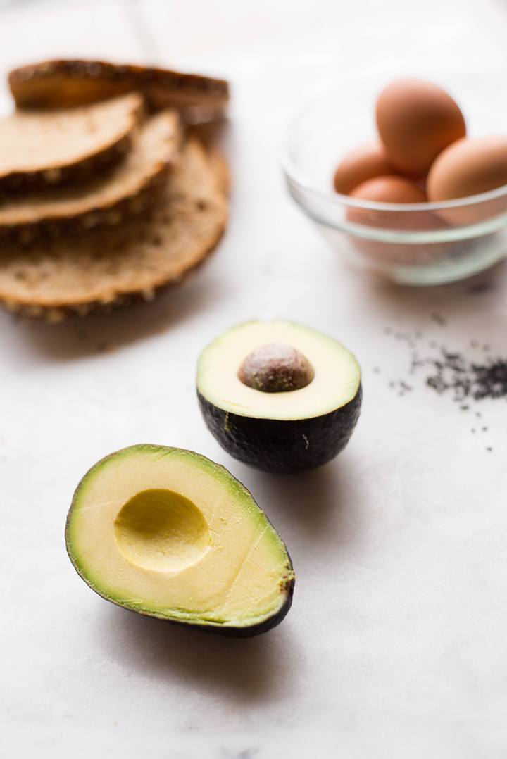 View of Perfect Avocado Toast ingredients including an avocado cut open, a few slices of whole grain bread, and a glass bowl containing eggs in the shell.