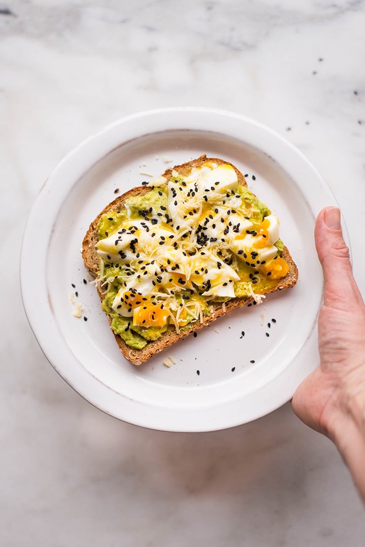 Overhead image of a hand holding a white plate with perfect avocado toast, topped with a soft boiled egg.