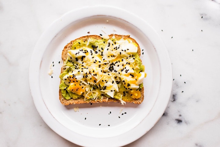 Overhead image of a slice of Perfect Avocado Toast on a white plate, with a soft boiled egg on the top of the avocado.