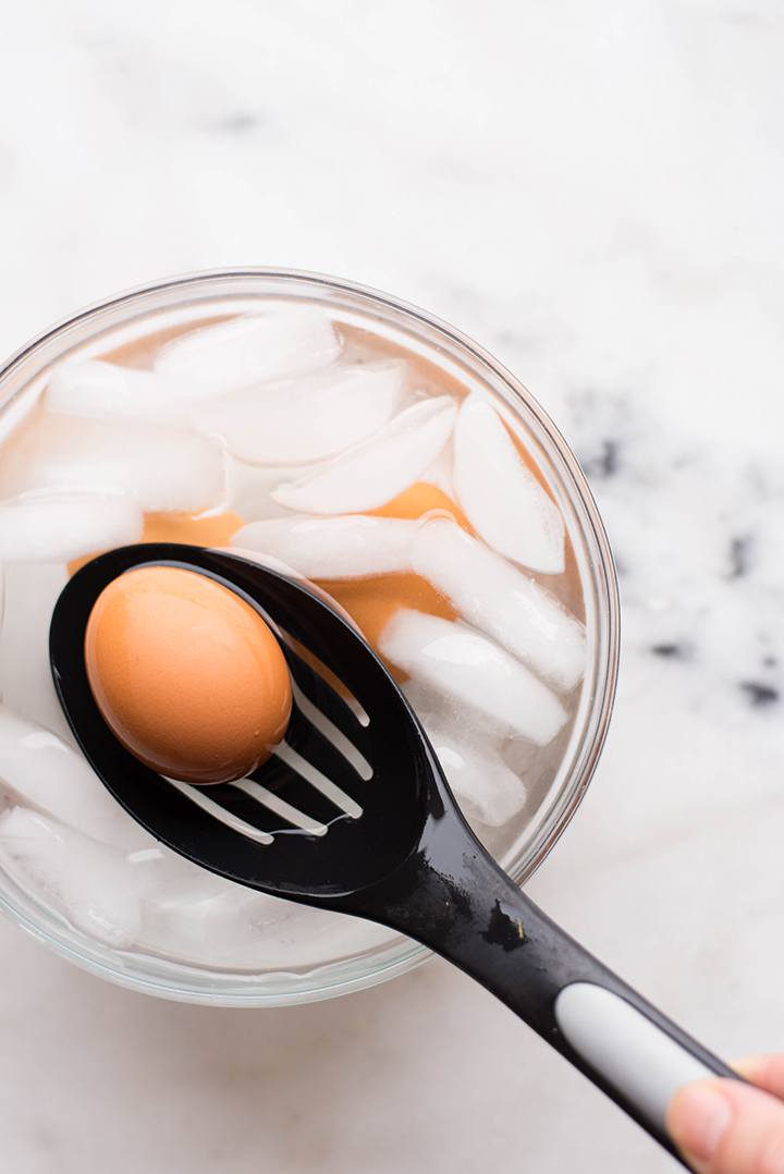 Overhead view of a soft boiled egg on a slotted spoon to be used in Perfect Avocado Toast, being lowered into a bowl of ice water.