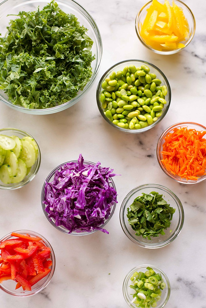 Overhead view of clear glass bowls with ingredients for Chopped Thai Salad with Peanut Dressing, including carrots, kale, edamame, and cucumbers.