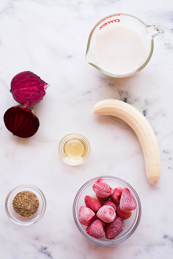 The ingredients required to make a pink power protein beet smoothie, including a raw beet, almond milk, frozen strawberries, banana, flaxseed meal, and raw honey.