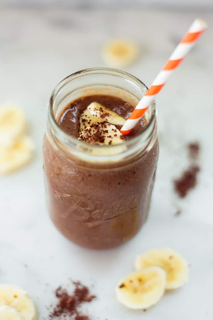 Overhead image of a mason jar containing chocolate banana protein smoothie, with a striped straw sticking out of the jar and banana slices around it.