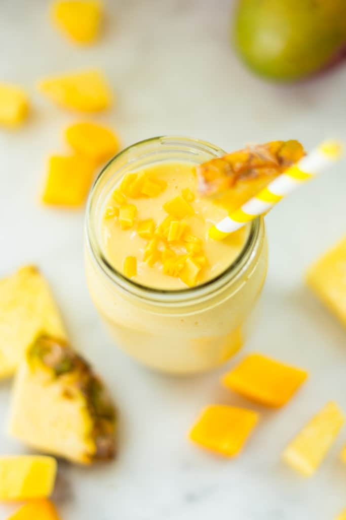Overhead view of the Vanilla Cupcake Smoothie in a mason jar with a straw in the jar and a piece of pineapple on the rim, and pineapple surrounding the jar.
