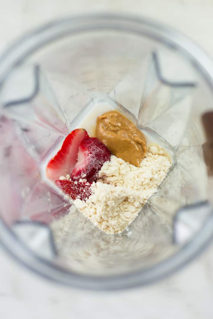 Overhead image of a Vitamix blender containing protein powder, peanut butter, and fresh strawberries ready to blend.