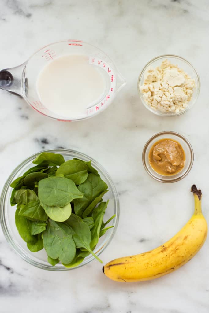 Overhead view of ingredients for the Green Machine Smoothie including baby spinach, banana, peanut butter, almond milk, and vanilla protein powder.