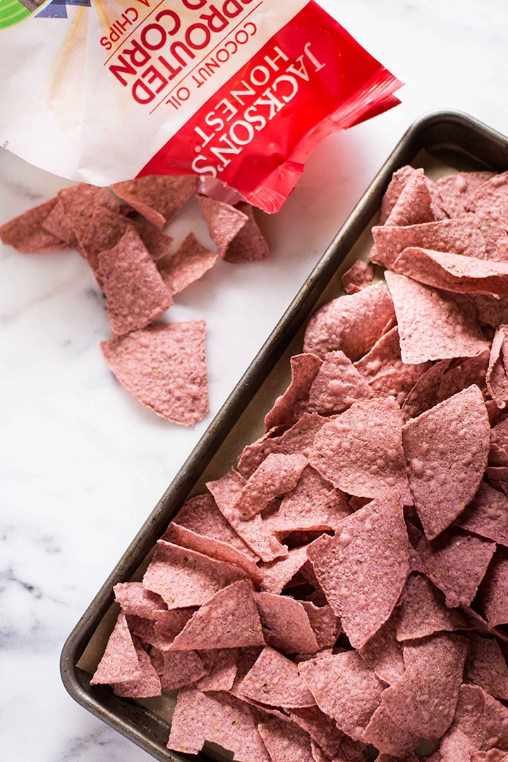 Bag of Jackson's Honest Sprouted Red Corn Tortilla Chips with tortilla chips coming out of the bag next to sheet pan lined with parchment paper and layered with more red corn tortilla chips