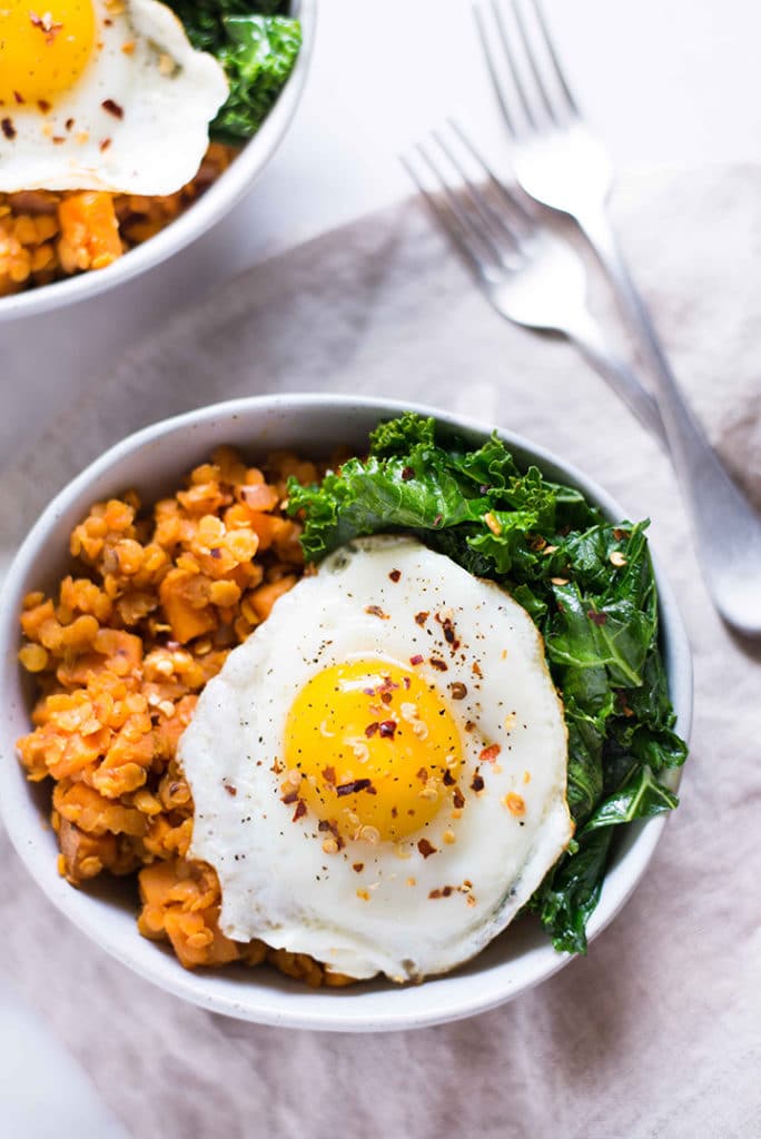 Overhead view of two bowls filled with sweet potato and lentil hash, garlic sauteed kale, and topped with fried egg with crushed red pepper flakes and black pepper sprinkled over the top.