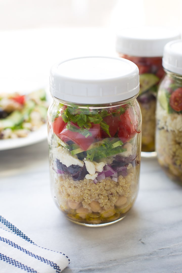 Close up side image of a mason jar containing chickpeas, quinoa, red onion, tomatoes, and cucumbers. Other mason jars and a bowl with ingredients are behind it.
