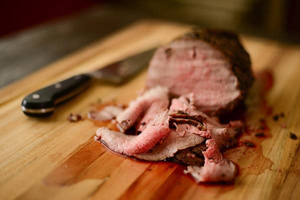 Close up view of freshly sliced roast beef on a wooden platter, with a knife laying beside it.