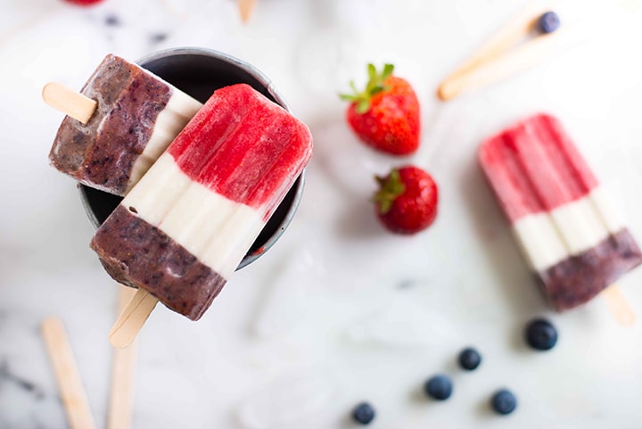 Overhead view of three red white and blue popsicles with fresh strawberries and blueberries on a cutting board.