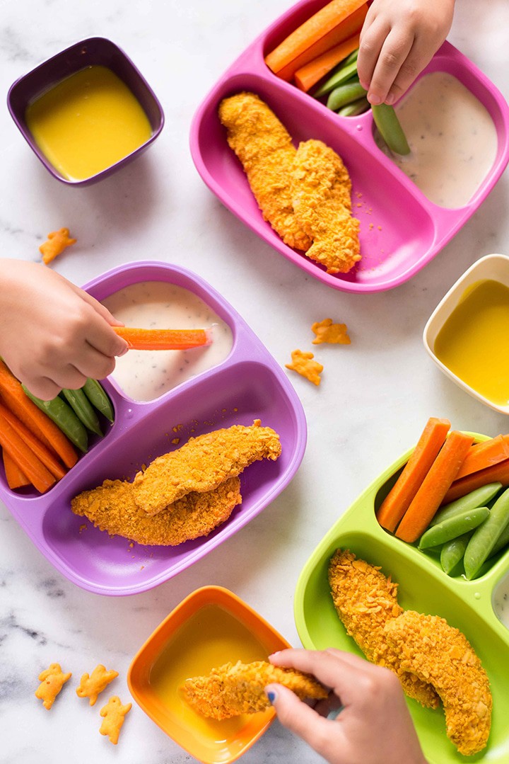 Close up overhead view of Kid-Friendly Cheddar Crusted Chicken Strips, showing two hands dipping the chicken and veggies.