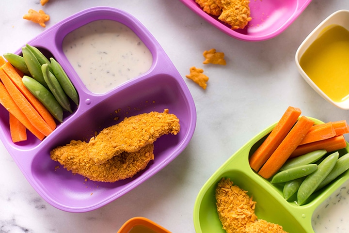 Close up photo of child's plate filled with cheddar-crusted chicken strips, carrot sticks, snap peas, and Annie’s Homegrown ranch dressing for a dip