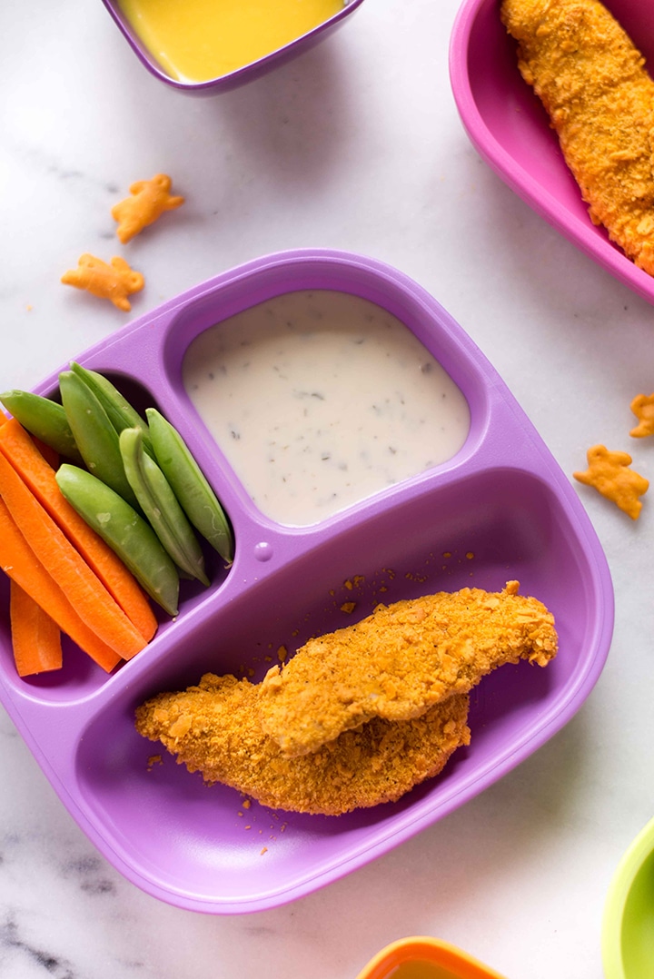 Two kid-friendly plates filled with cheddar-crusted chicken strips, carrot sticks, snap peas, and Annie’s Homegrown ranch dressing for a dip next to bowl with honey mustard dipping sauce for the chicken strips