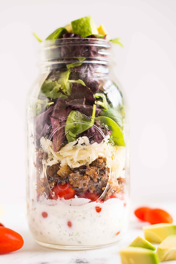 Quart size mason jar filled with taco salad recipe with creamy cilantro lime dressing, along with grape tomatoes, taco meat, shredded jack cheese, and mixed baby greens.