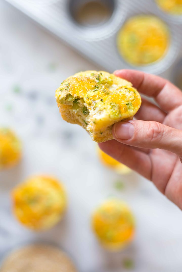 Hand holding baked healthy egg muffin cup that contains quinoa, broccoli, and eggs and has a bite out of it.
