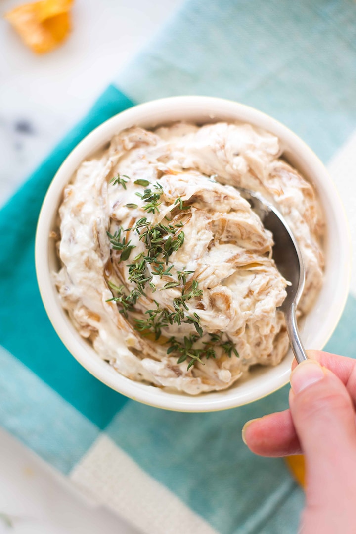 Overhead view of a bowl of caramelized onion dip topped with fresh thyme leaves with a hand taking a spoonful of the dip.