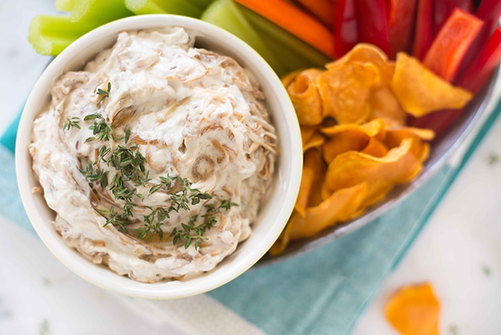 Horizontal image of the caramelized onion dip that is ready to be eaten as an appetizer with fresh veggies and baked sweet potato chips.