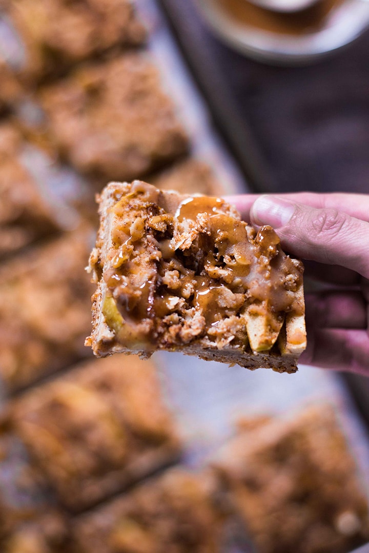 Hand holding a single apple pie bar to show the texture. In the background is the sheet pan lined with the remaining apple pie bars and the salted caramel sauce.