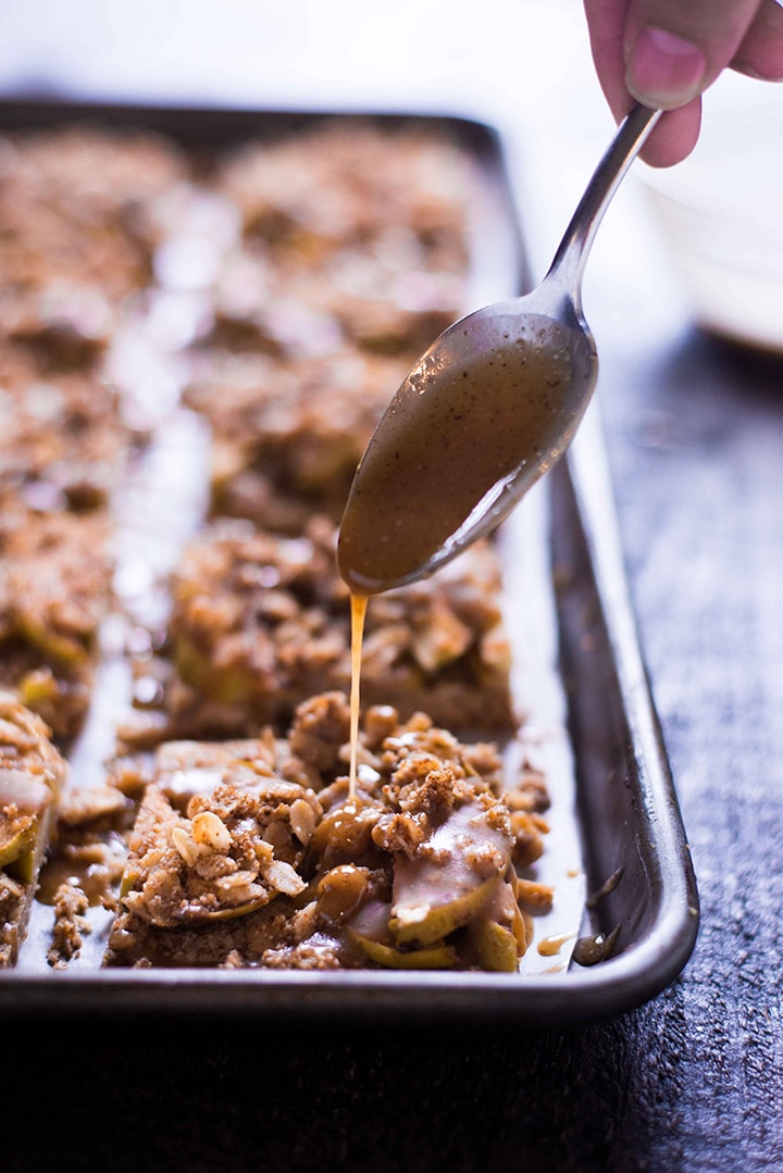 Spoon that is drizzling the salted caramel sauce over the apple pie bars that have been baked and divided into squares.