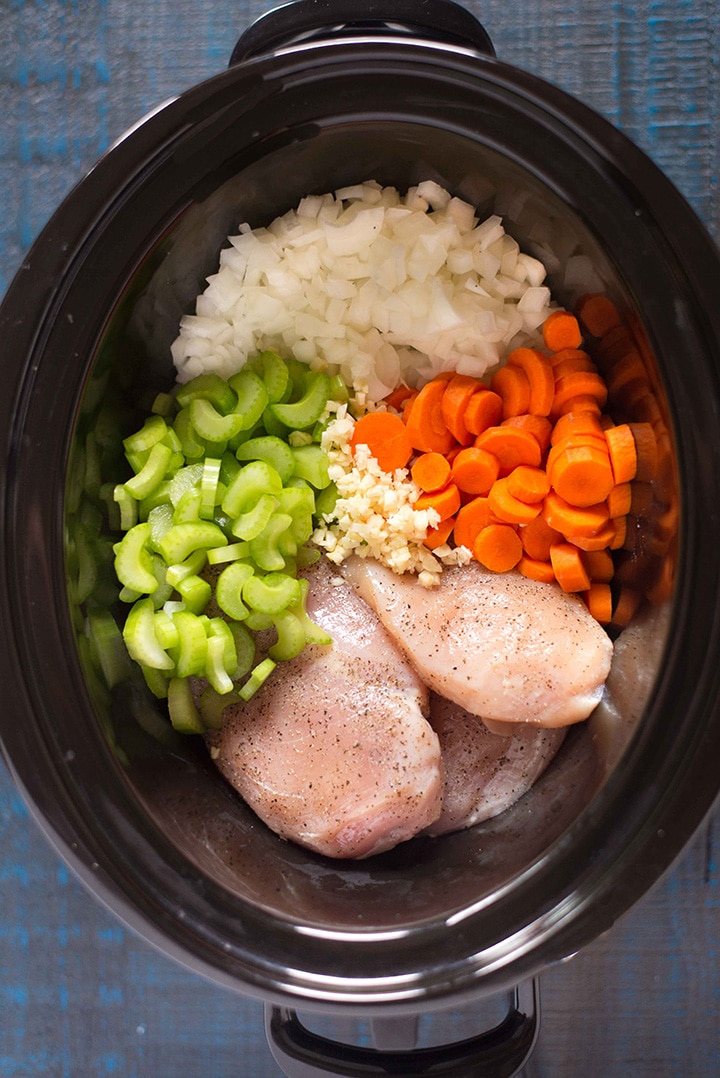 Bowl of the slow cooker filled with the boneless, skinless chicken breasts that are seasoned with sea salt and black pepper, sliced carrots, sliced celery, yellow onion, and garlic, ready to be cooked on Low in the slow cooker to make the chicken noodle soup.