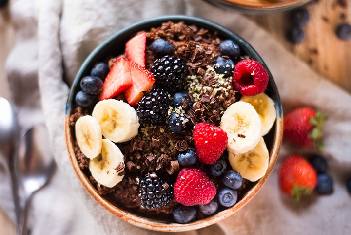 Horizontal image of the Chocolate Quinoa Breakfast Bowl which is sitting next to fresh strawberries, blueberries, and chocolate shavings.