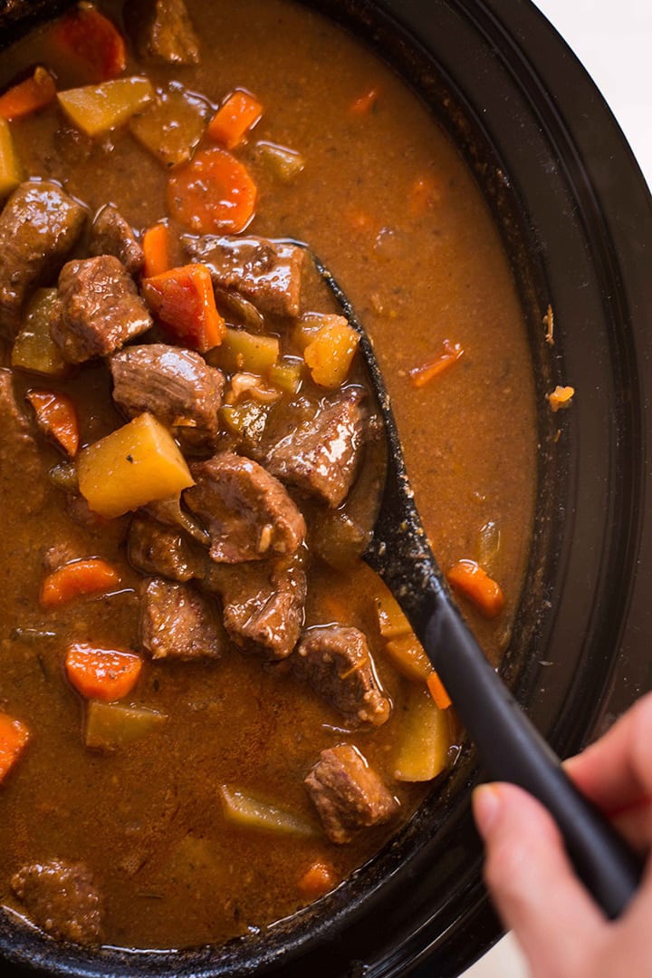 Close up overhead image of a slow cooker bowl with beef stew, with a hand holding a spoon and stirring the stew.