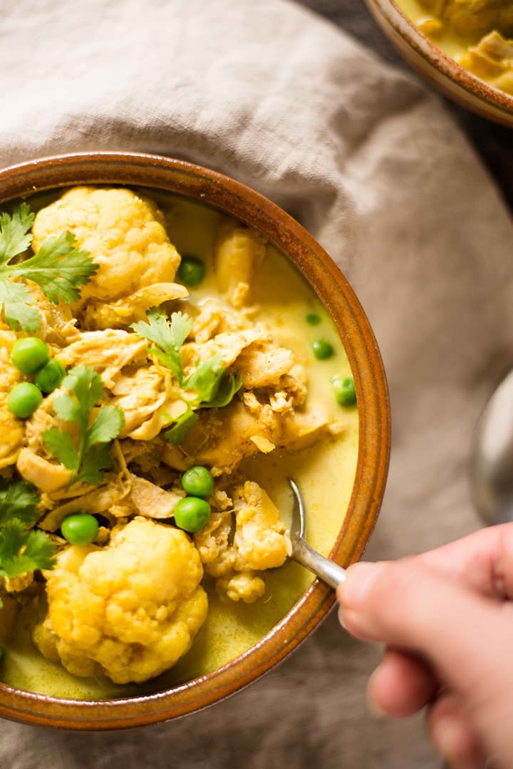 Overhead image of a hand with spoon scooping out some of the slow cooker chicken cauliflower curry from the ceramic bowl.
