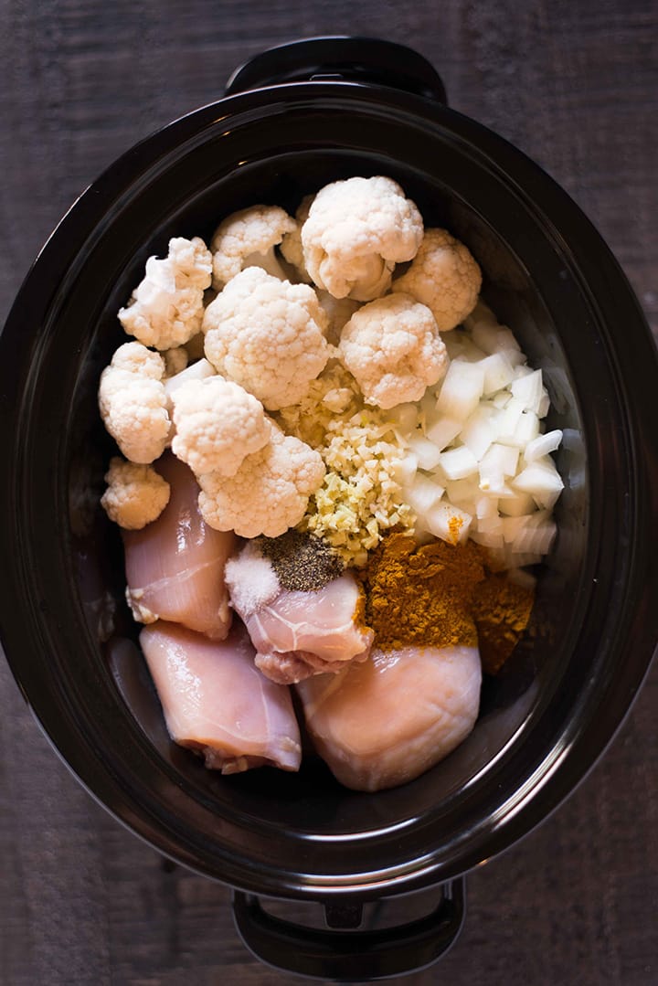Overhead of the slow cooker with all the ingredients for the slow cooker chicken cauliflower curry recipe, including chicken thighs, cauliflower florets, garlic, ginger, yellow onion, curry powder, sea salt, and black pepper.