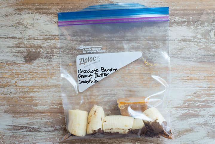 An overhead image of a smoothie freezer pack lying on the kitchen counter with the ingredients for the Chocolate Banana Peanut Butter Smoothie including chopped banana, peanut butter and chocolate whey protein powder.