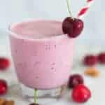 A side image of a glass of Cherry Vanilla Almond Smoothie with pitted cherries, plain greek yogurt, whole almonds, vanilla extract, almond extract, raw honey and almond milk decorated with a single fresh cherry.
