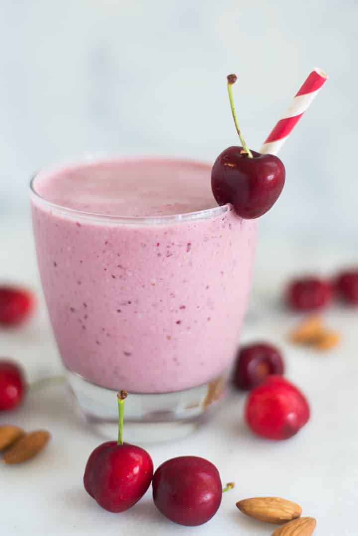 A side image of a glass of Cherry Vanilla Almond Smoothie with pitted cherries, plain greek yogurt, whole almonds, vanilla extract, almond extract, raw honey and almond milk decorated with a single fresh cherry.