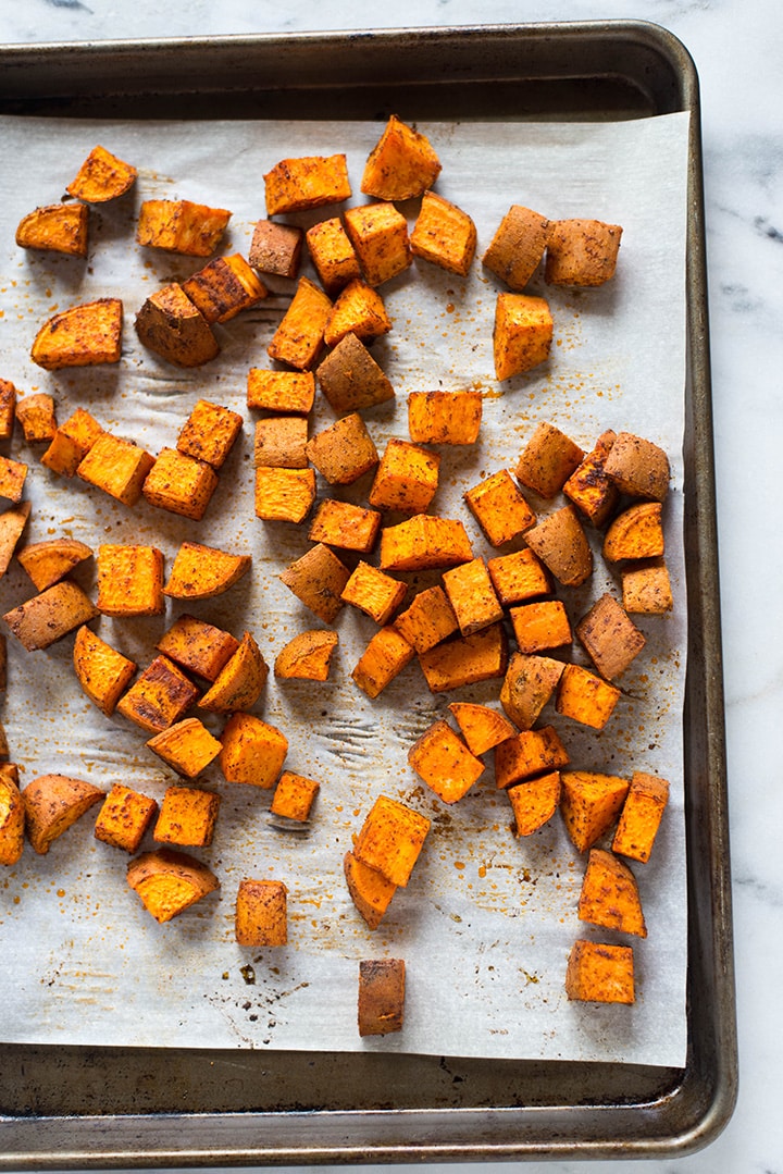 Overhead view of a rimmed baking sheet that is lined with parchment paper and is topped with the seasoned sweet potatoes that will be roasted to use in the chicken protein bowl.
