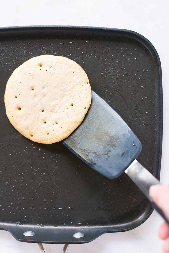 Overhead view of a griddle that is greased with coconut oil and a spatula is flipping a cooked banana oat banana pancake to the other side to show how long to cook a pancake before flipping.